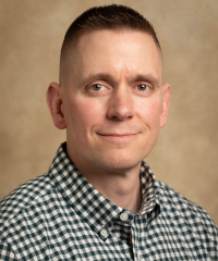 Headshot of Dr. Nicholas Shallcross in a white and green checkered shirt infront of a brown background